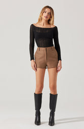 A woman is wearing a black, long-sleeved, open-knit top that is slightly sheer and has a wide boat neckline. She pairs it with high-waisted, camel-colored shorts with a front zipper and side pockets. She completes the outfit with tall, black, knee-high boots.